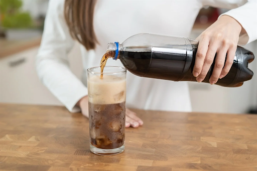 A person pouring a dark soft drink from a plastic bottle into a glass on a wooden table