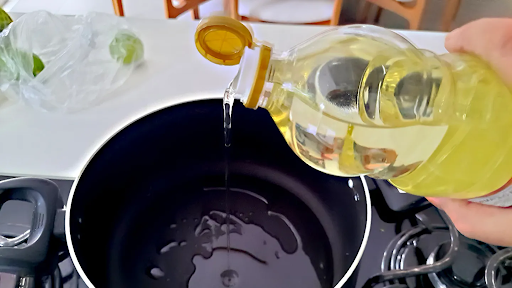 A hand pours light yellow cooking oil from a clear plastic bottle into a large black pot on a gas stove, with a white countertop and kitchen items blurred in the background.