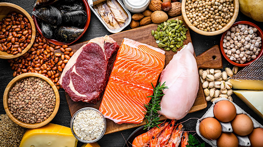 Top-down view of a variety of high-protein foods, including salmon, steak, chicken breast, eggs, shrimp, nuts, seeds, beans, and legumes arranged on a dark surface, showcasing both animal and plant-based protein sources.