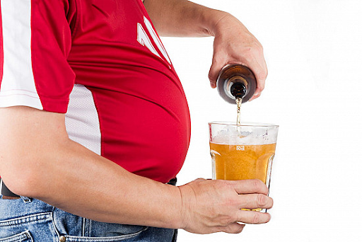 A close-up of a man sitting outdoors holding two large plastic cups of beer, highlighting excess belly fat associated with increased heart disease risk.